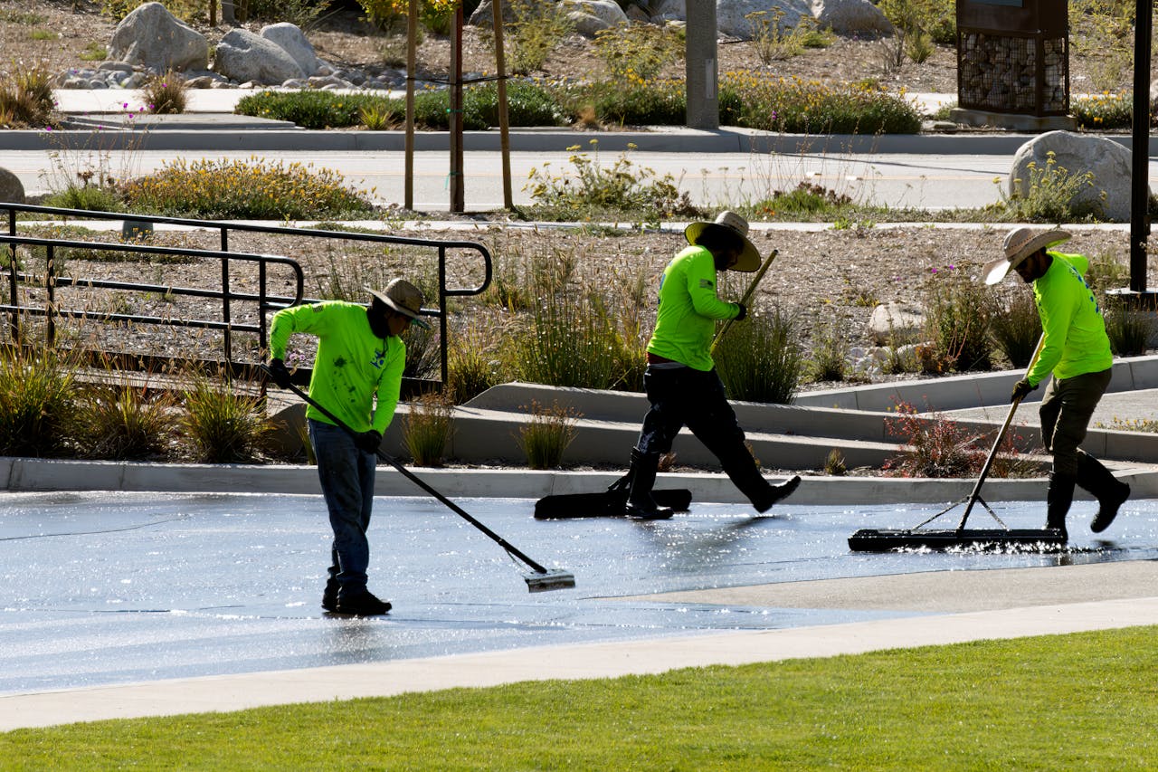 Crew of workers in bright uniforms cleaning a park walkway with brooms under sunlight.